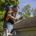 A roofing contractor stands on a ladder and inspects a roof while speaking on a cellular telephone.