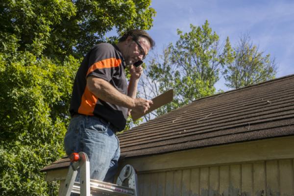 A roofing contractor stands on a ladder and inspects a roof while speaking on a cellular telephone.