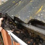 A hand cleans debris out of a gutter as a roof hangs over it.