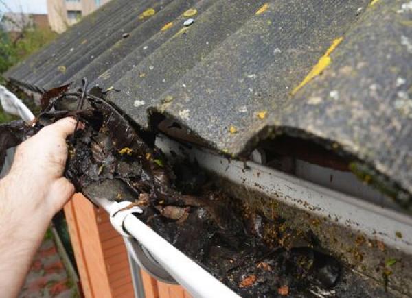 A hand cleans debris out of a gutter as a roof hangs over it.